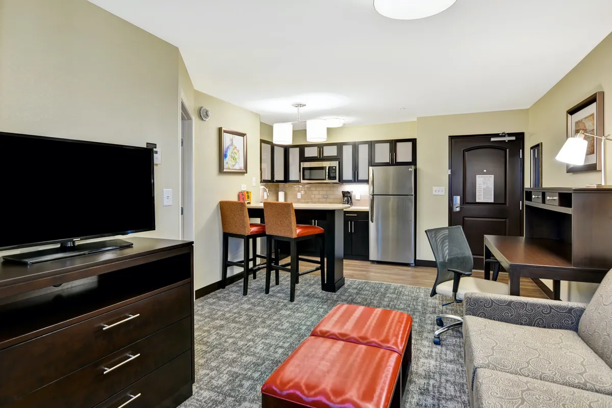 A stylish, sunlit condo interior featuring a modern kitchen with bar stools, a cozy living area with a plush sofa and ottoman, and a wall-mounted television. The space is designed for comfort, with functional areas for dining, relaxing, or working, ideal for couples or solo travelers. The setting feels inviting and practical, perfect for unwinding after a day exploring Mount Juliet or Nashville.