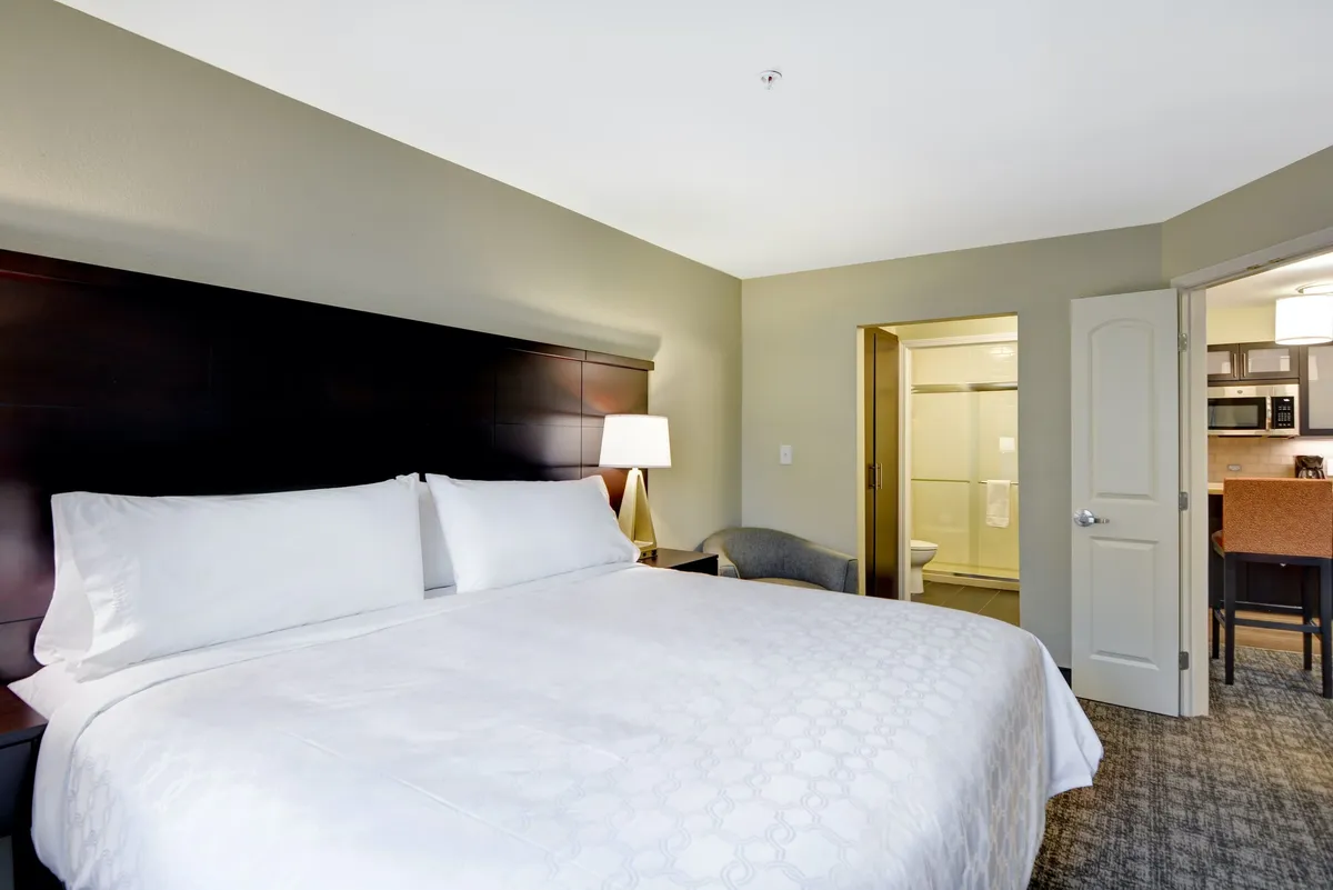 This image features a spacious bedroom with a large bed dressed in white linens, a nightstand with a lamp for evening reading, and an armchair. The scene includes doorways leading to the bathroom and kitchenette area, contributing to the sense of a self-contained, private suite. The overall mood is calm and comfortable, emphasizing restful and cozy evenings.