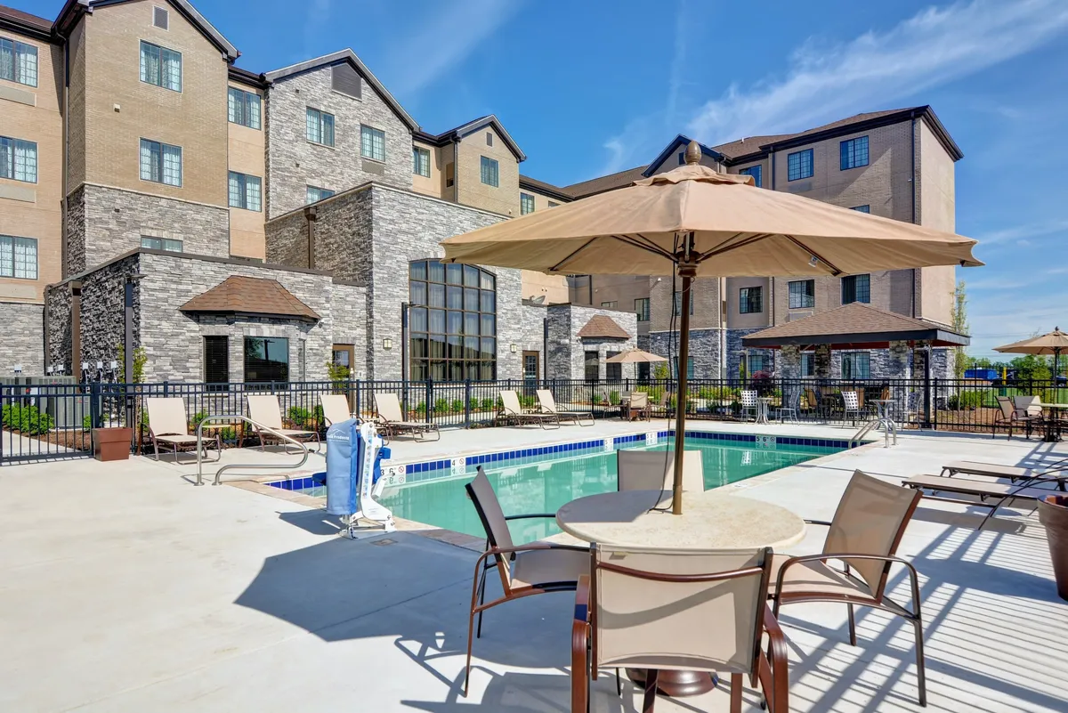 The scene features a well-maintained swimming pool with umbrellas and lounge chairs, adjacent to the hotel building. The mood is leisurely, capturing sunny afternoons and potential evening gatherings by the pool.