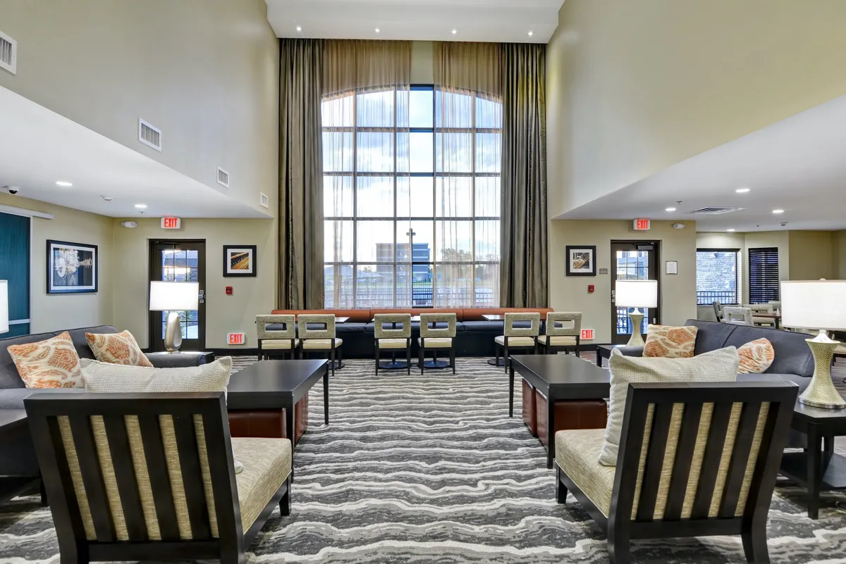 The image displays a bright and welcoming lounge area featuring large windows framed by sheer curtains, stylish seating arrangements with colorful cushions, and modern light fixtures. The view from the windows suggests a connection to the outside neighborhood, making the room feel airy and integrated with the local setting.