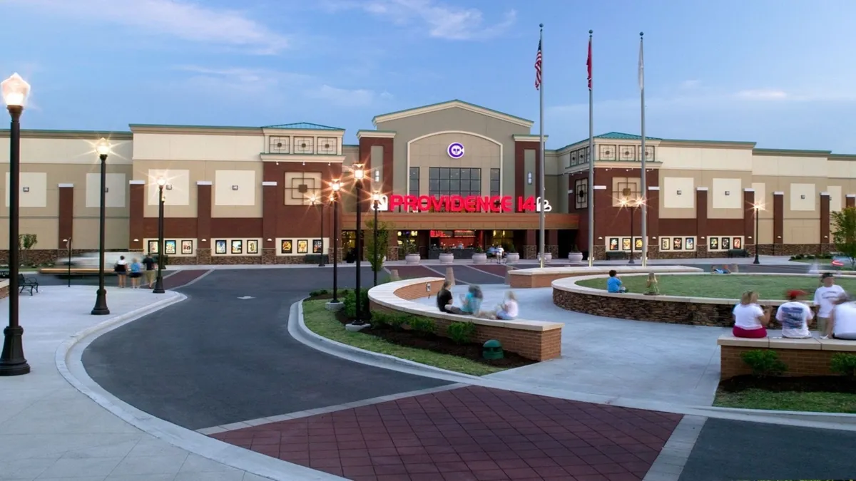 This exterior image shows the entrance to a theater, surrounded by a green lawn and illuminated by attractive light posts. The space feels lively, suggesting an area where families might gather to watch movies, socialize, or participate in community events.