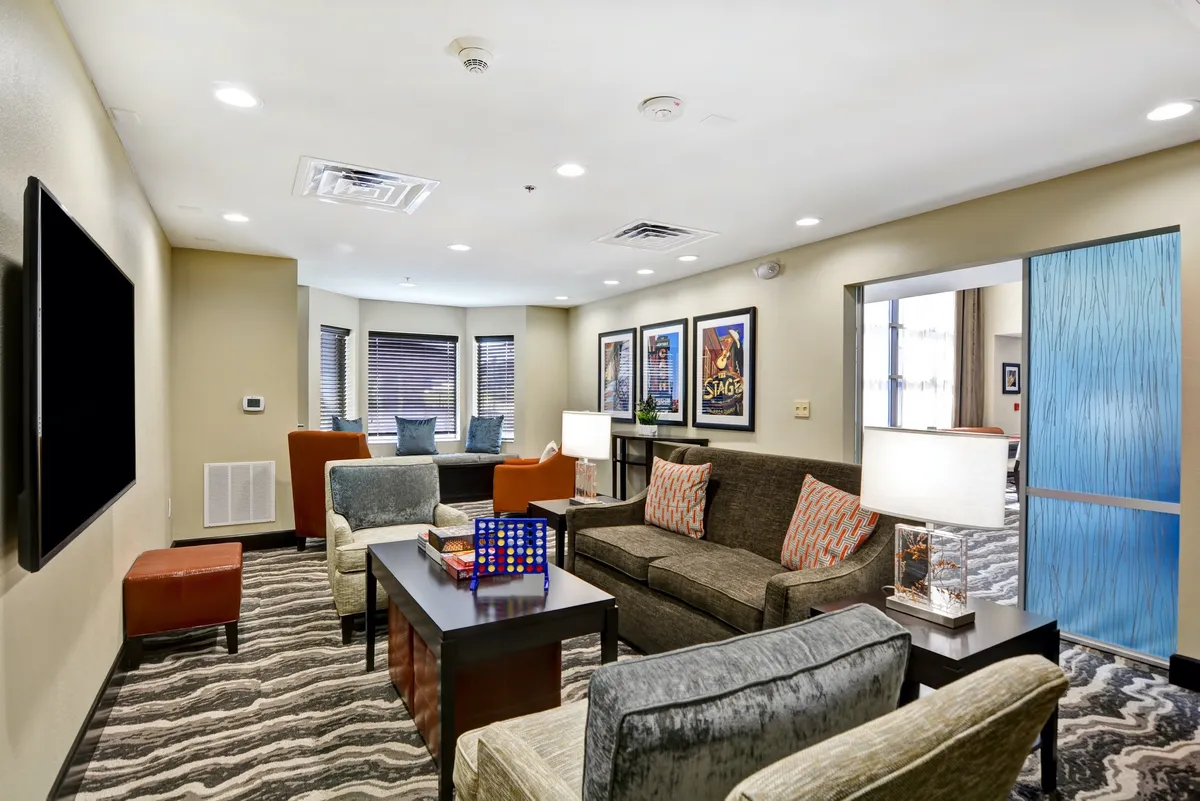 The living area is showcased with a comfortable sofa with patterned cushions, a coffee table set up for board games, framed artwork on the wall, and a large flat-screen TV. The overall atmosphere is casual and welcoming, perfect for relaxing, watching TV, or working in comfort.