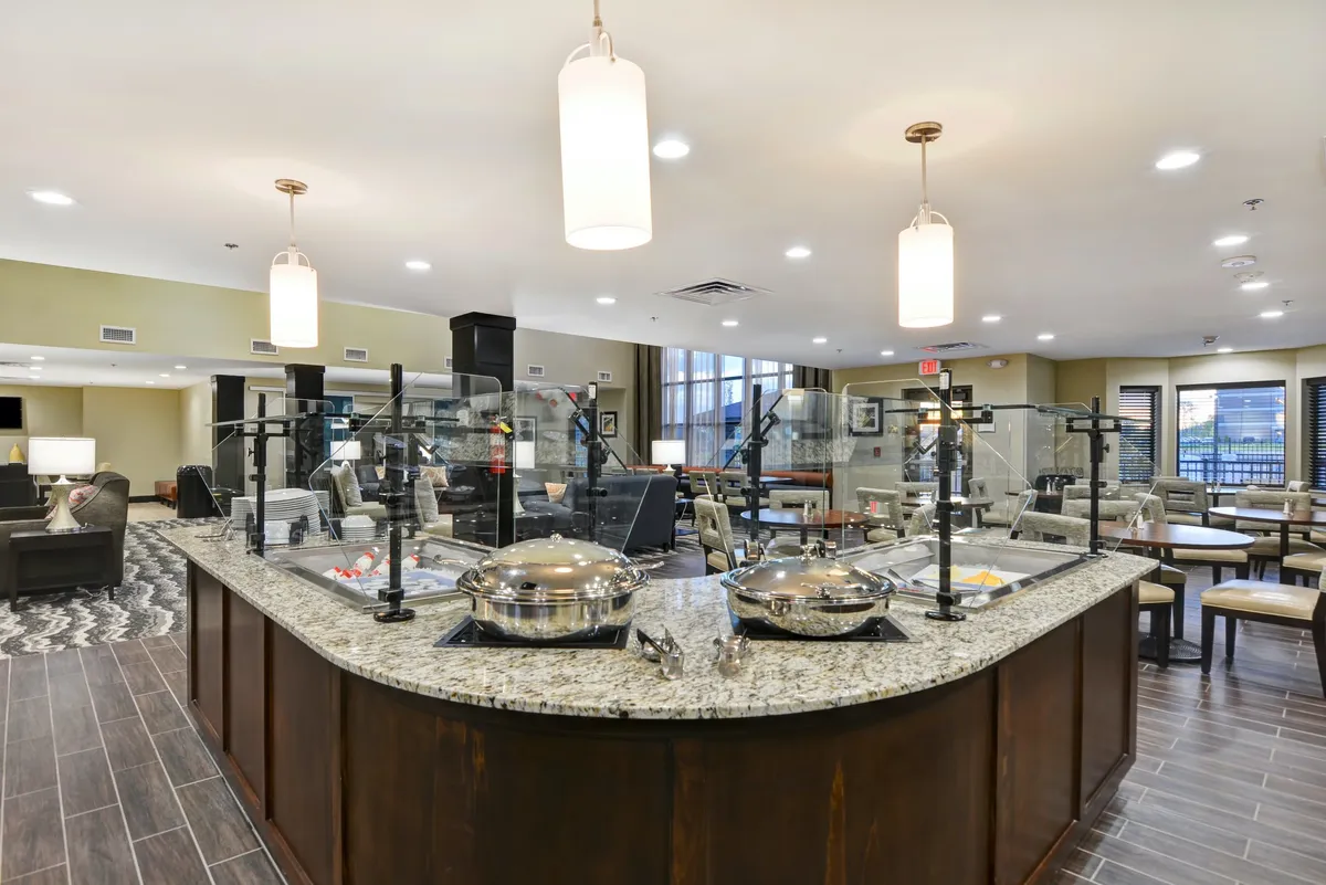 A spacious communal area with a granite countertop, serving dishes set up for meals, and a seating area near large windows. The scene conveys a sense of shared dining and communal enjoyment, suitable for both family dinners and casual meals.
