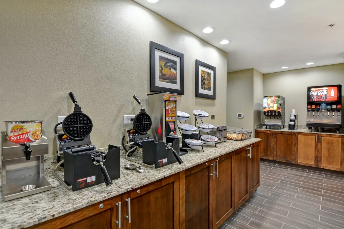 This image focuses on the breakfast area, equipped with waffle makers, a coffee machine, and a beverage dispenser. The setup encourages self-service and the enjoyment of a complimentary breakfast.