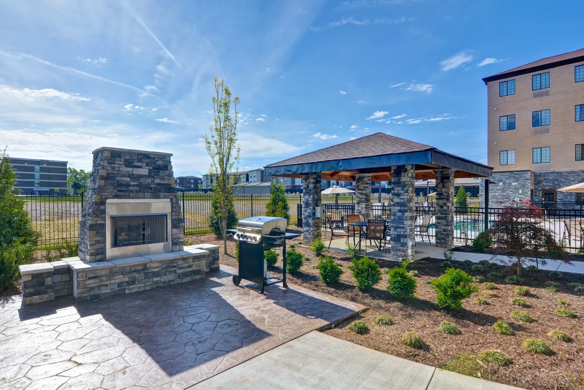 This exterior image showcases a beautifully landscaped garden with a stone fireplace, a grill, and a gazebo. The setting is ideal for outdoor cooking, relaxing by the fire, or enjoying meals al fresco. The lush plantings and communal spaces evoke a sense of relaxation after a busy day.